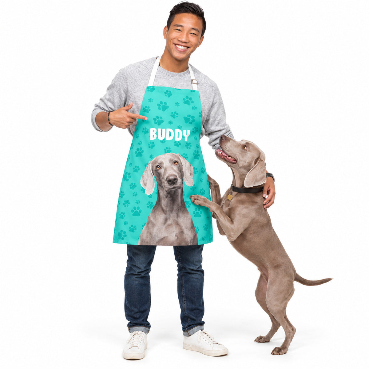 A man wearing a custom printed teal apron with a Weimaraner dog graphic and the name 'BUDDY' in white, standing next to a real Weimaraner dog on a white studio background.